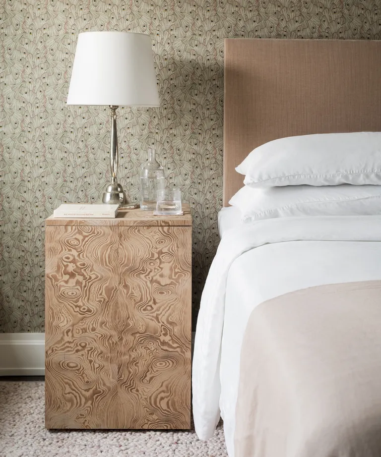 A bedroom with a wooden side table, white lamp, neutral wallpapered walls and a pink textured carpet