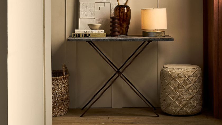 A warm, softly lit hallway with layered beige walls featuring subtle arch detailing. A brass wall sconce and matching table lamp cast a gentle glow over a console table styled with books, vases, and dried flowers.
