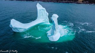 The phallus-shaped iceberg floats near a bench-shaped berg in the water, near the coast.