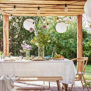 Dining table under a wooden pergola in the garden, with cream linen table cloth and festoon lights strung overhead