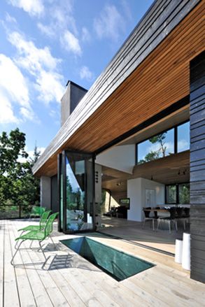 Close up exterior view of the outdoor terrace at Trekronekabin during the day. The terrace features wooden decking, green chairs and glass balustrades. The glass doors to the house are open offering a partial view of the living area and the kitchen and dining area