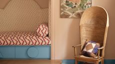 Quirky peach fuzz bedroom with bed alcove and wicker egg chair. There are mismatched patterns including zig zags on bed, dots on alcove headboard and geometric patterns on the egg chair cushion. The under bed storage and baseboards are a chalky blue