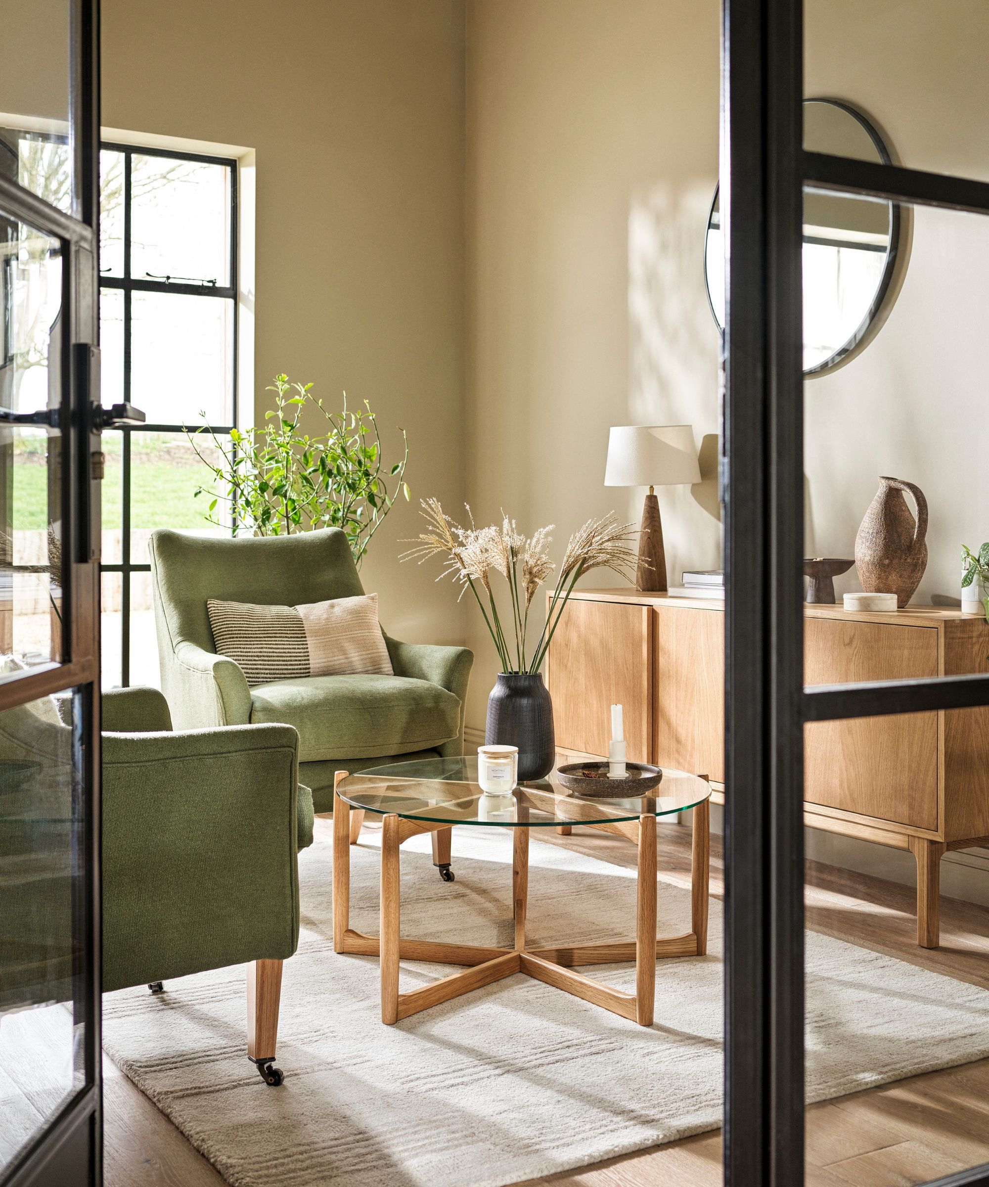 A calm sandy-toned sitting room seen through black steel doors, with sage green armchairs, a round glass coffee table on oak legs, oak sideboard and dried grasses in a black vase.