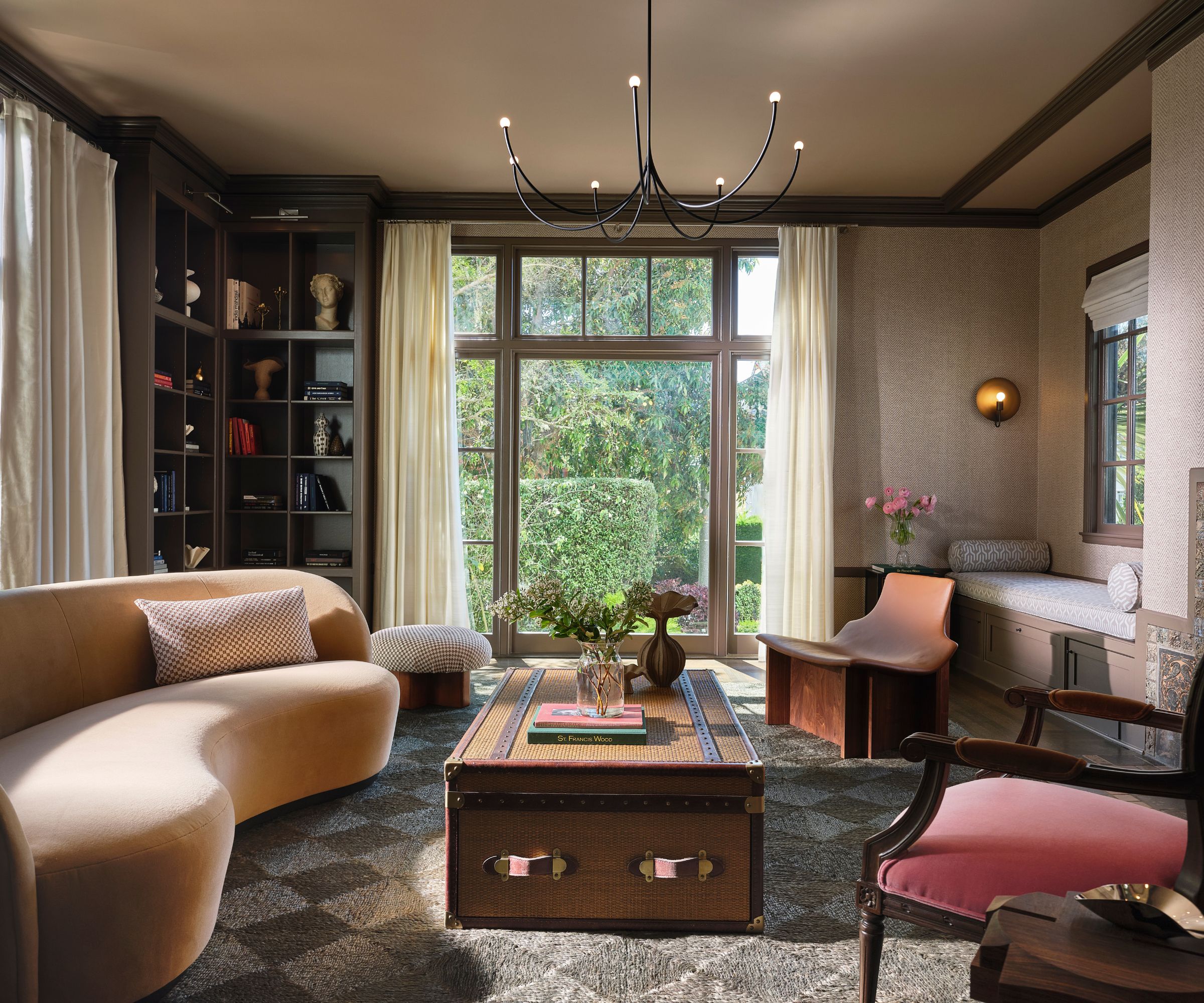 A living room with taupe wallpapered walls, dark brown trim, a large floor-to-ceiling window with cream curtains, a corner of built-ins, a beige sculptural sofa, modern armchairs, and a chest coffee table