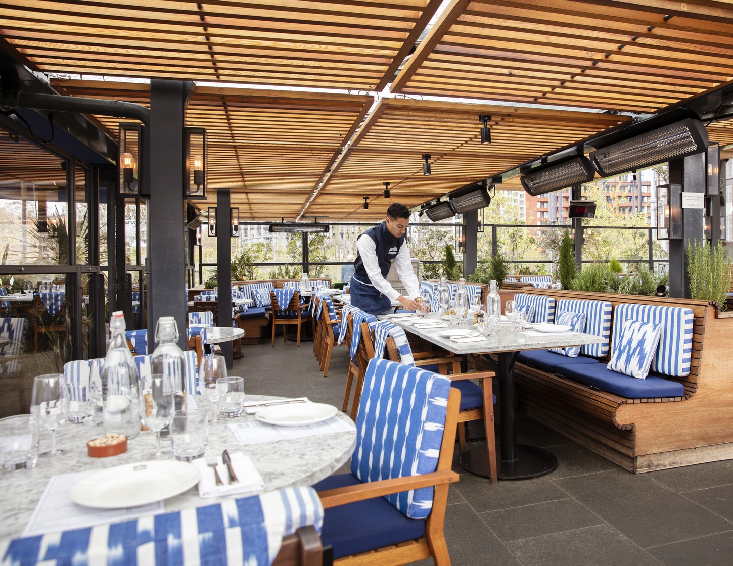 Restaurant terrace with a stone floors, wood seating with blue and white cushions, a wooden awning and a lone waiter