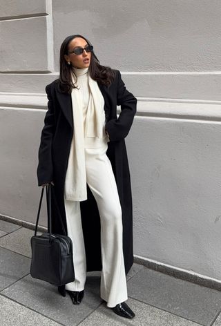 The winter capsule wardrobe item, ankle boots, is shown in a photo of a woman standing outside wearing black sunglasses with a long black coat layered over a white scarf sweater with matching knit pants, black ankle boots, and a black bag