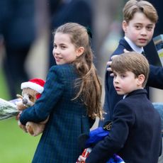 Princess Charlotte wearing a blue plaid coat holding a stuffed sloth with a Santa hat turning around and smiling next to Prince Louis, who is clutching a mickey doll, and Prince George who is turned around in a blue suit