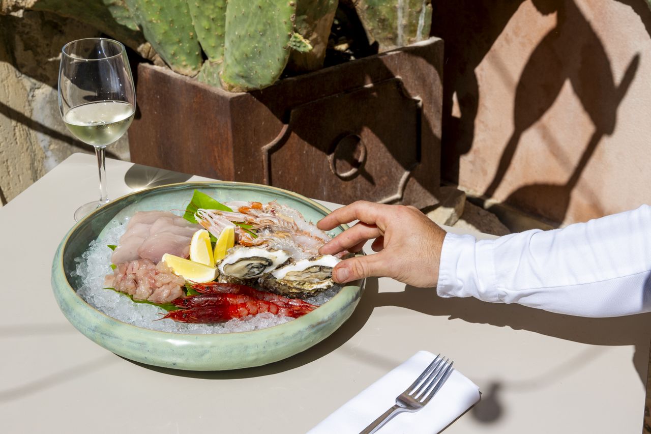 A man's hand is photographed while reaching for an oyster served on ice along with other crudo seafood at a table in the sun.