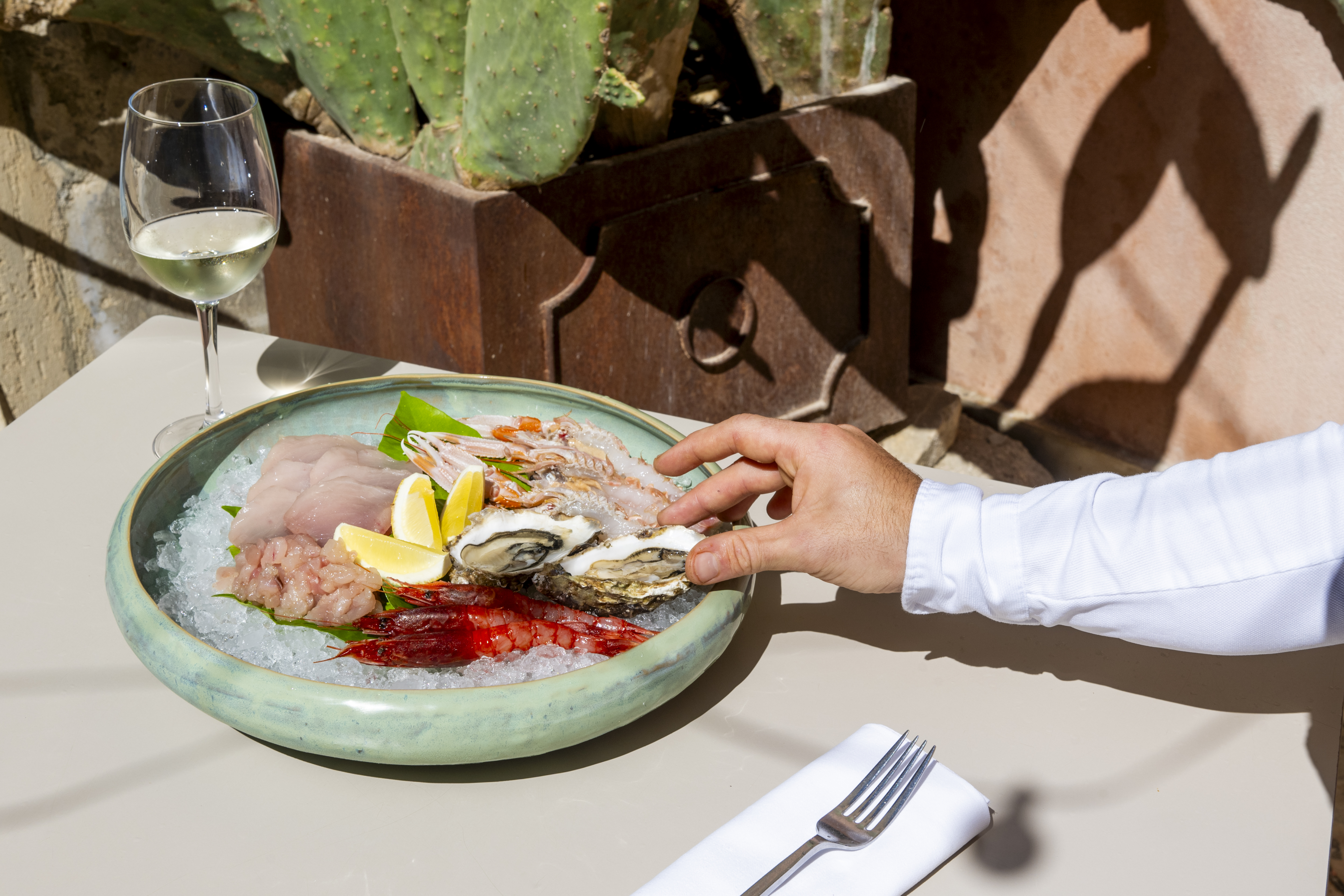 A man's hand is photographed while reaching for an oyster served on ice along with other crudo seafood at a table in the sun.