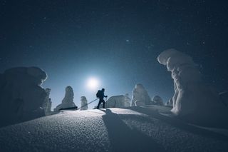 Person walking over snow with sled, silhouetted by the moon with stars and some ice around