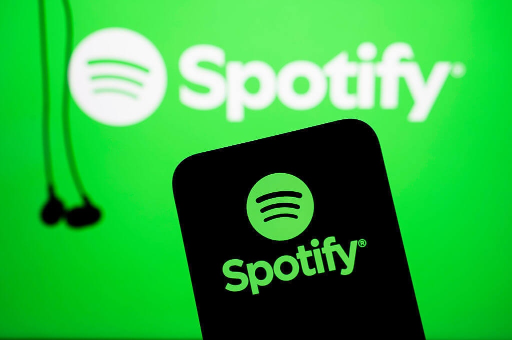 A phone showing the Spotify logo in green with a pair of earbuds next to the phone against a green background that has the Spotify logo in white