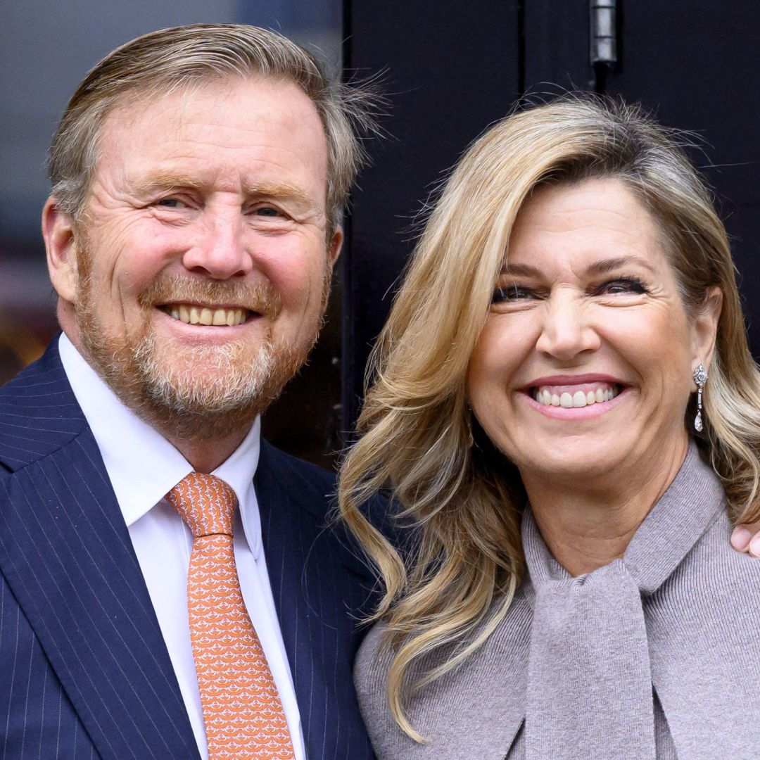Queen Maxima wearing a gray coat and King Willem-Alexander wearing a blue suit and orange tie smiling 