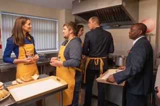 Kate Middleton and Prince William wearing yellow aprons working in a kitchen with staff