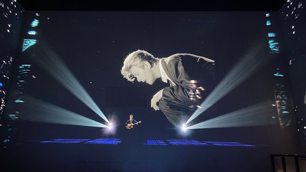 A projected image of David Bowie performing on stage while a huge black and white version of himself crouches down next to him in the exhibition David Bowie: You're Not Alone at Lightroom, London.