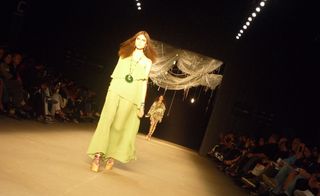 Model on runway wearing a layered, pale green dress with a green pendant necklace