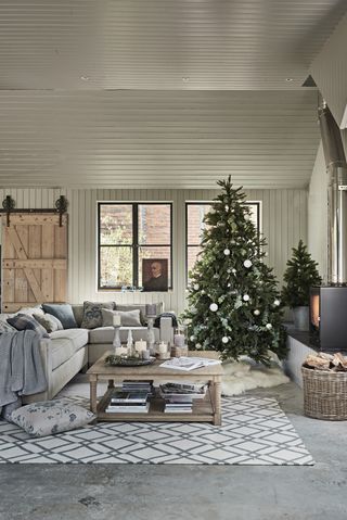 A cozy Christmas living room by Neptune with concrete grey floor, geometric motif white and grey rug, Christmas tree, L-shaped grey sofa, and white shiplap walls and ceiling
