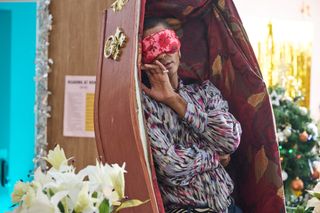 Stephen (Layton Williams) lies upright inside a luxuriously-upholstered coffin, positioned vertically. He is wearing a floral eye mask.