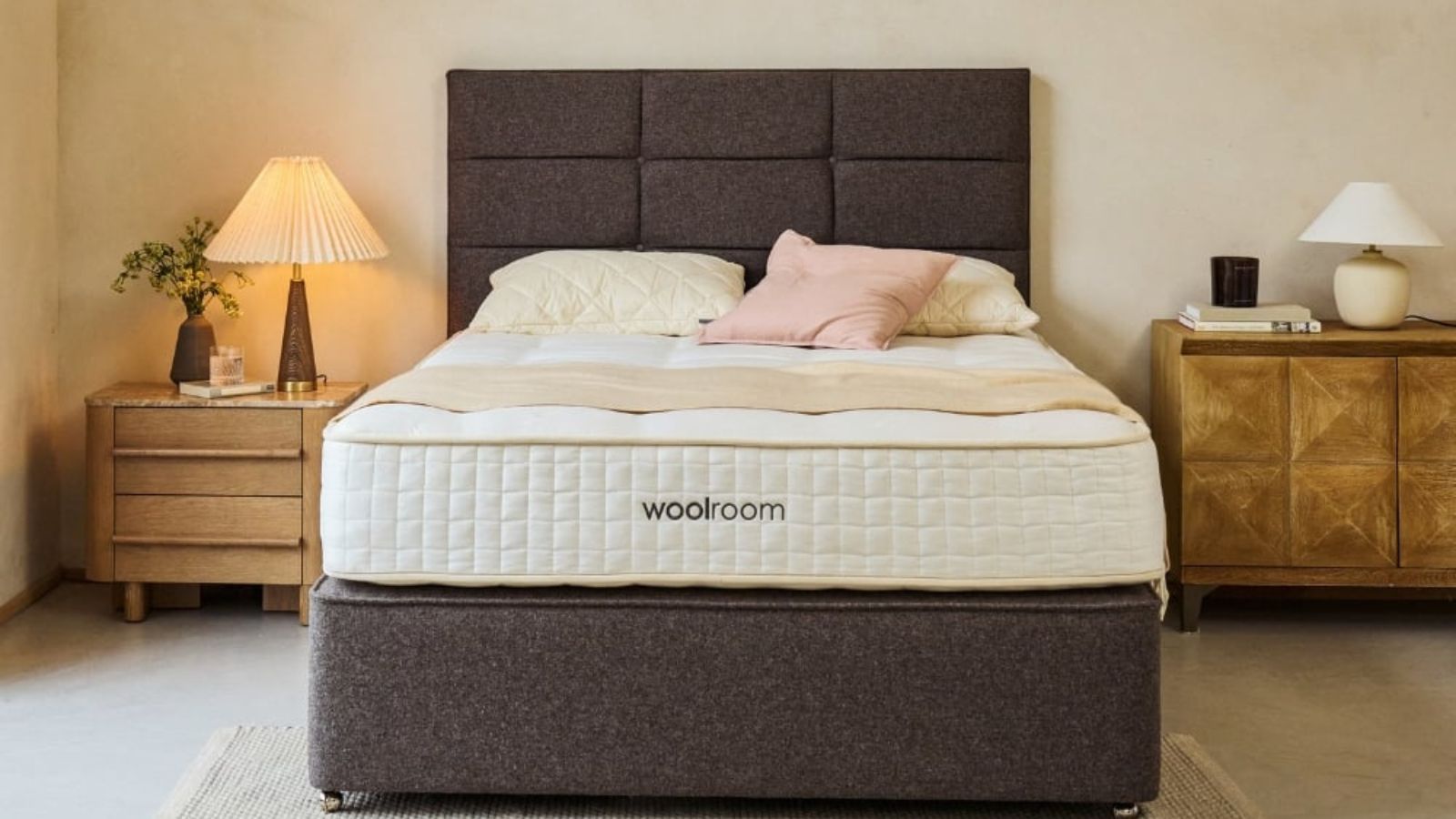 A Woolroom mattress on a gray bed frame against cream walls; a nightstand and a lamp either side of the bed. 
