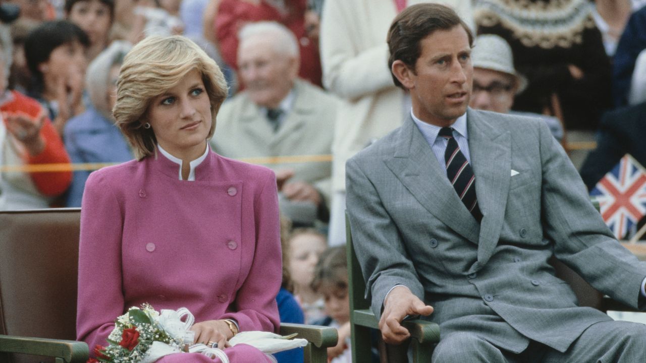 Prince Charles wearing a gray suit and Princess Diana in a pink suit holding flowers sitting in chairs next to each other with a crowd behind them