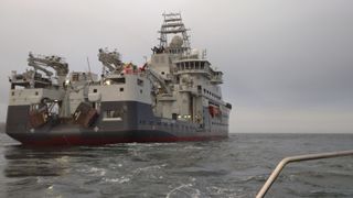 A large boat is shown on the left-hand side of the image as it crosses the sea. The edge of a metal bar from another vessel is shown in the bottom, right-hand corner of the image.