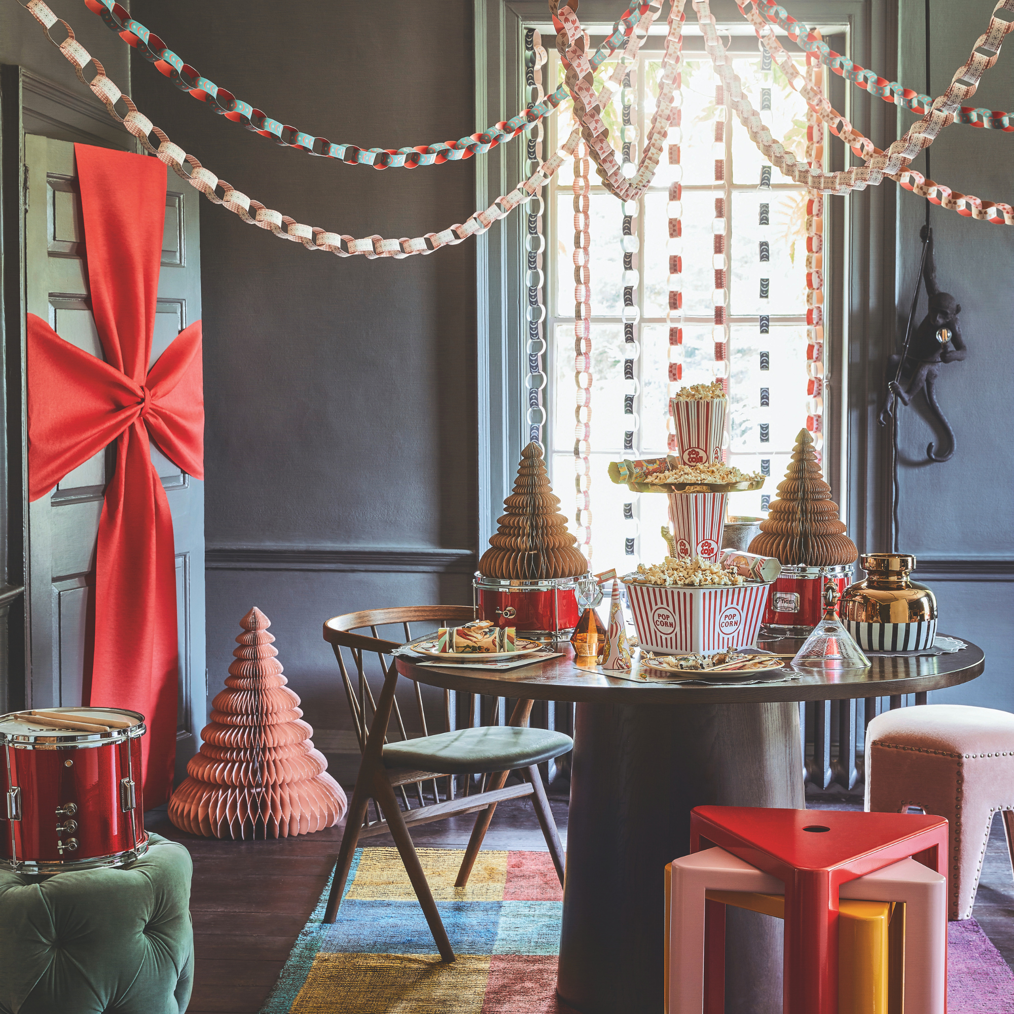 Dining room decorated in blue and pale green with ribbons of paper chains, circular table surrounded by stools and chairs on a multicoloured rug. Green door wrapped with red ribbon. A circus themed Christmas decoration scheme.