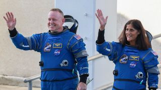 two astronauts in blue space suits wave and smile