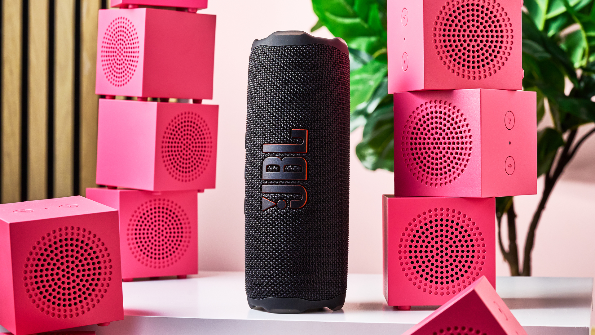 The JBL Flip 7 standing on its end surrounded by piles of pink IKEA Kallsup speakers.
