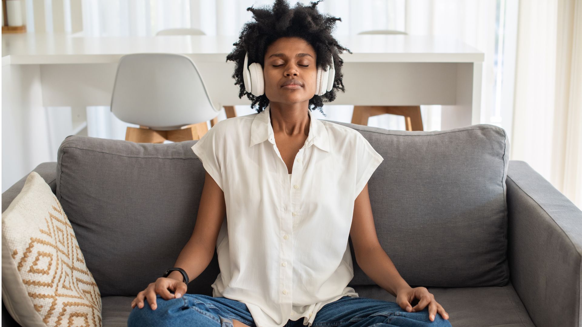 A woman sits cross-legged on a couch with her hands resting on her knees. She has her eyes closed and is wearing headphones.