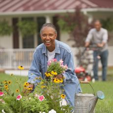 smiling woman planting flowers in the front yard while a man mows the lawn