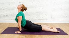 A woman performs a cobra stretch on a yoga mat. Her legs are straight with thighs facing the floor, while her torso is held upright by her straightened arms. Behind her we see a whitewashed brick wall