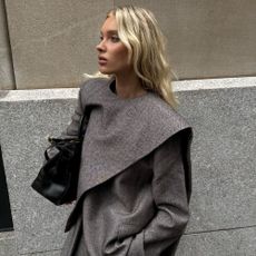 Elsa Hosk wears a grey scarf jacket with matching grey trousers in New York. She holds a black bag and wears black pointed-toe boots.
