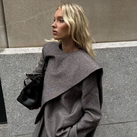 Elsa Hosk wears a grey scarf jacket with matching grey trousers in New York. She holds a black bag and wears black pointed-toe boots.
