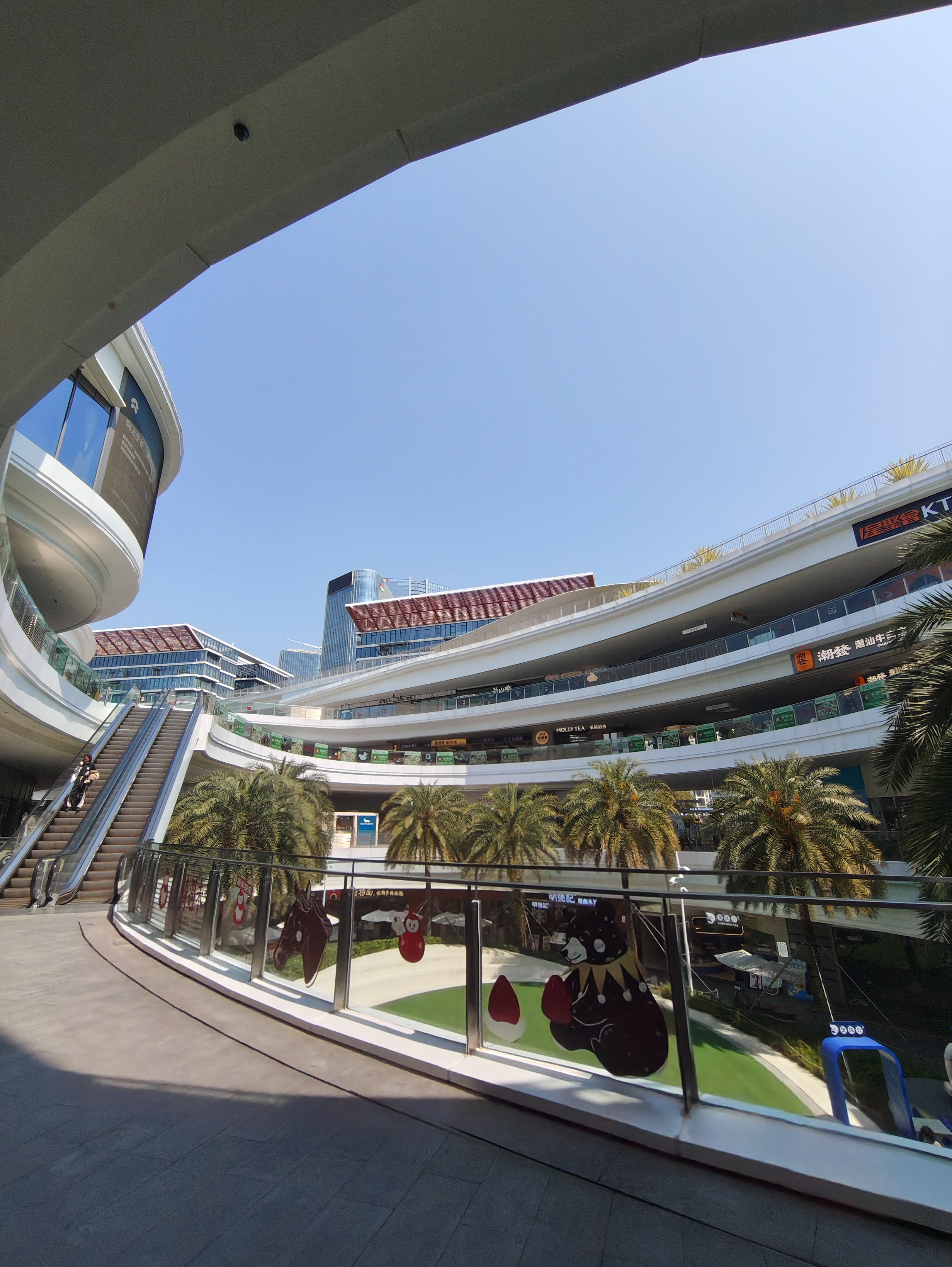 A wide-angle shot of a multi-story, modern outdoor shopping mall with white, curved balconies and glass railings. Escalators lead up the side of the building on the left. Several palm trees are planted in the central courtyard, and decorative stickers of cartoon characters are applied to the glass partitions in the foreground.