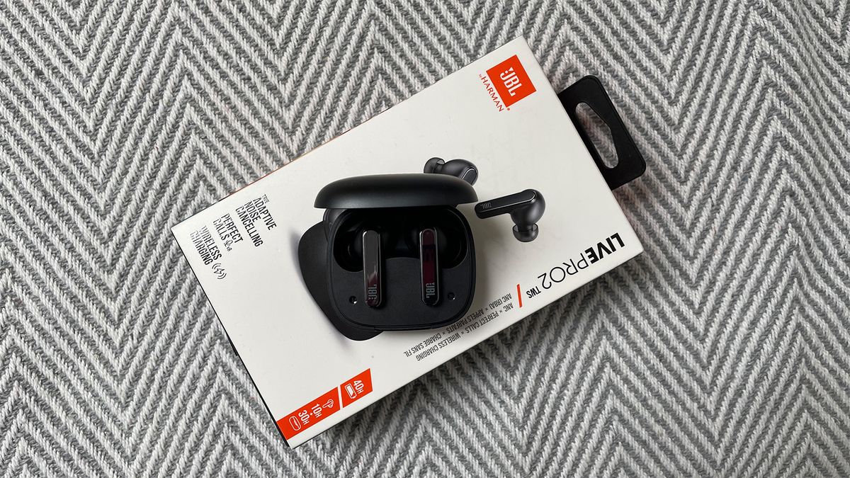 A top-down shot of a pair of black JBL Live Pro 2 TWS in-ear headphones inside the charging case with the lid open. They are placed on top of the box for the headphones, which is positioned on a fabric with a grey and white zigzag pattern.