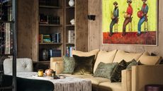 a burl wood clad sitting area with built in shelving, a beige couch and a painting of cowboys on the wall