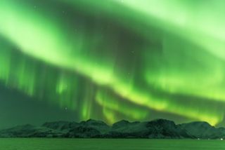 northern lights in Norway appearing as vivid green bands of light across the sky. With mountains and water below.