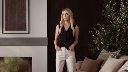 Interior designer Amber Lewis stands in a modern living room wearing a black vest top and her hands in her white trousers' pockets next to a white wall with a piece of brown art above the fireplace