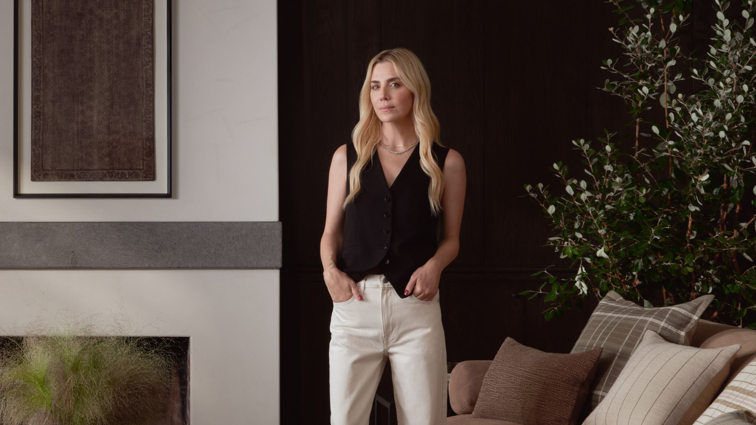 Interior designer Amber Lewis stands in a modern living room wearing a black vest top and her hands in her white trousers' pockets next to a white wall with a piece of brown art above the fireplace