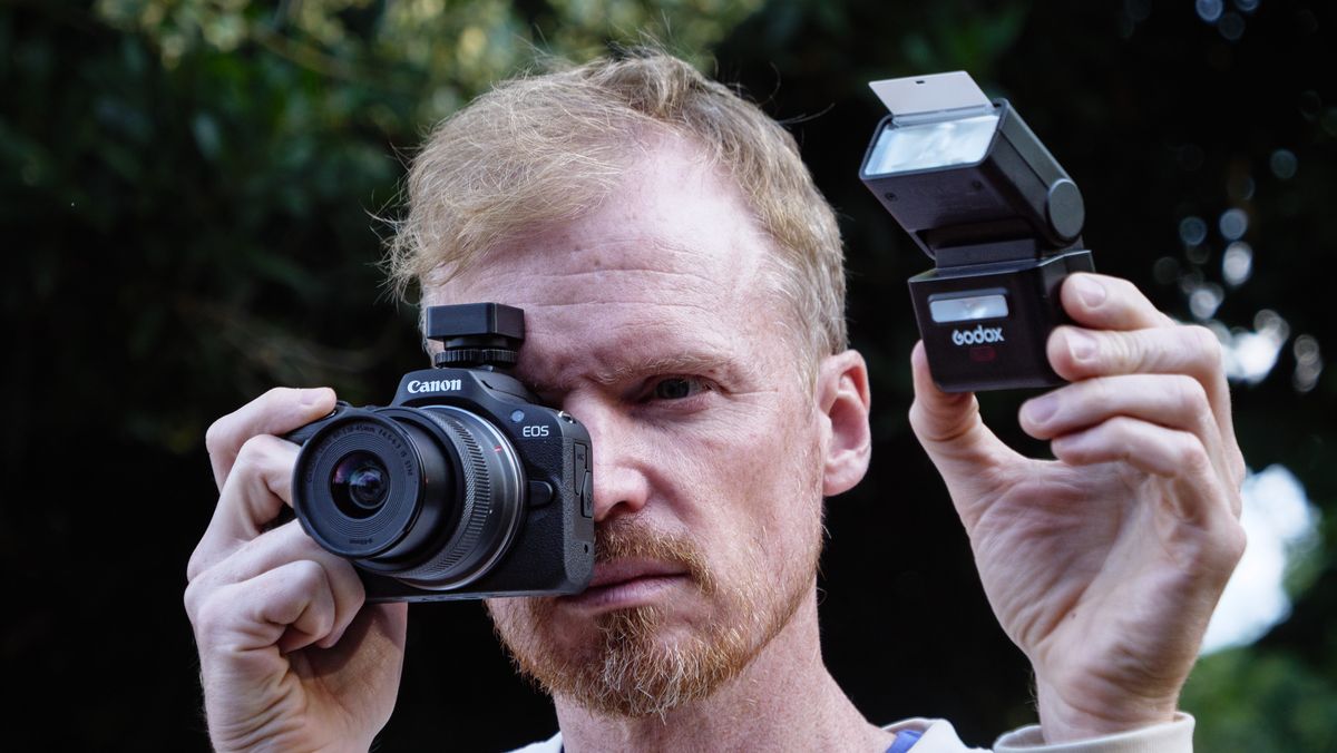 Photographer and TechRadar's Cameras Editor Tim Coleman holding a Canon camera up to his eye, there's a Godox iT32 camera flash alongside to the camera