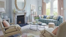 A pastel living room with blue patterned wallpaper, a white marble mantelpiece with circular mirror above, and large windows to the right with pastel pink curtains. In the foreground are two cream upholstered chairs, and in the center of the room is a circular coffee table with pink flowers on it, with a blue plaid sofa behind. On the floor is a white velvet rug.