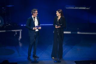 King Frederik and Queen Mary standing on stage in front of microphones lit by blue lights