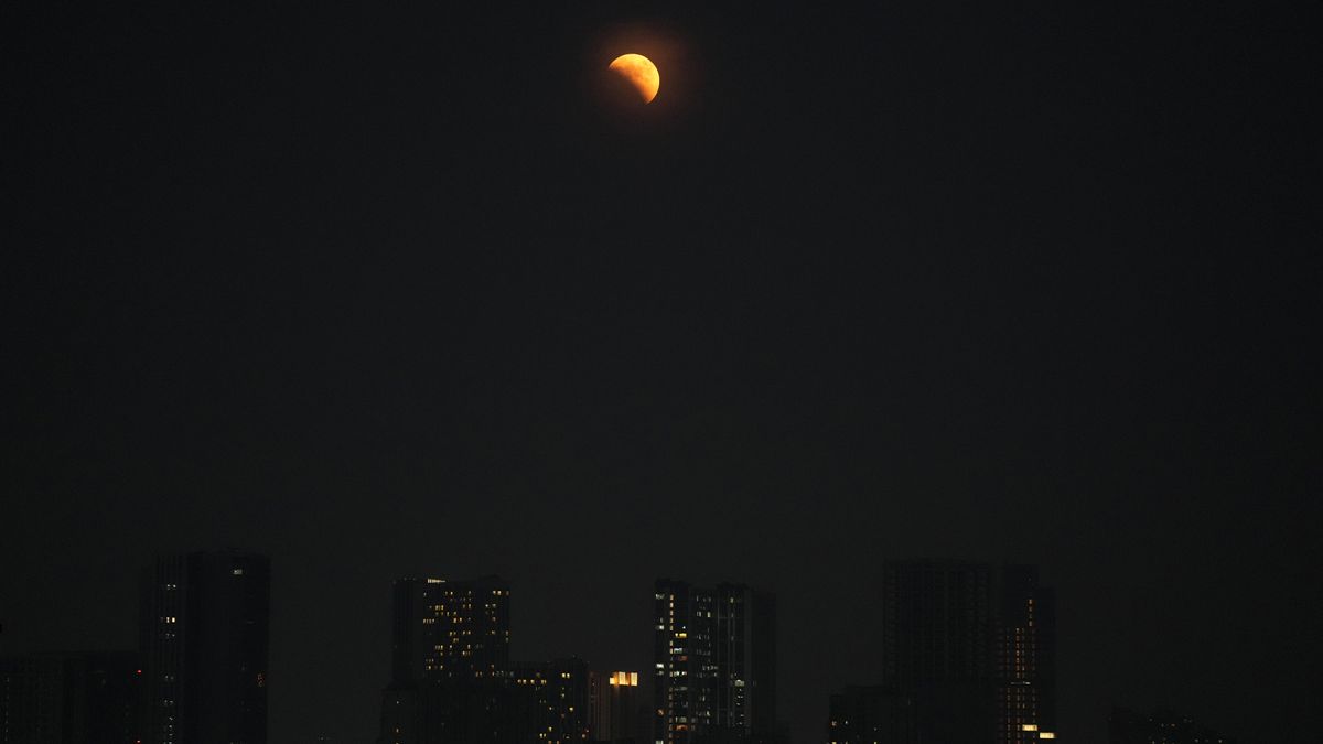 Partially lit orange moon with a city skyline below.
