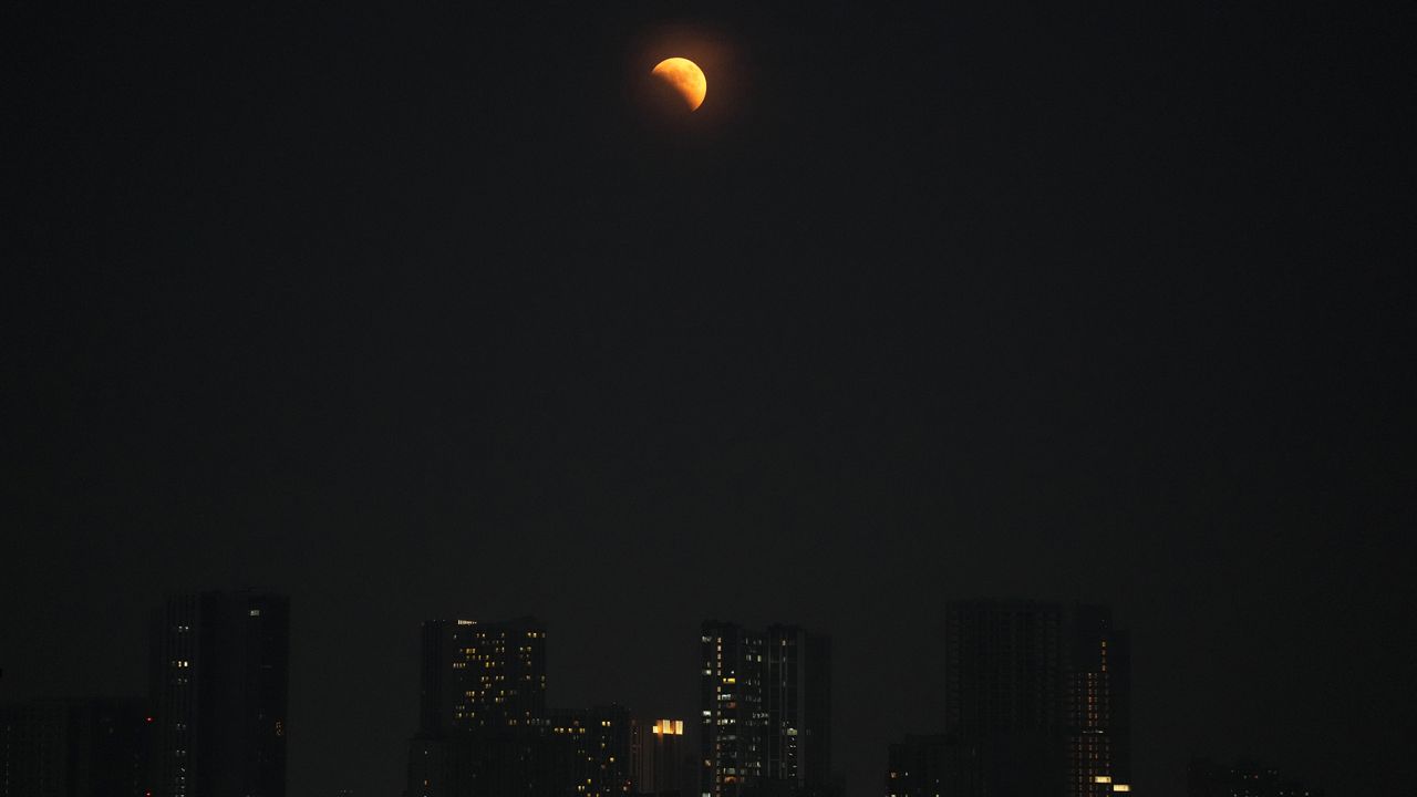 Partially lit orange moon with a city skyline below.