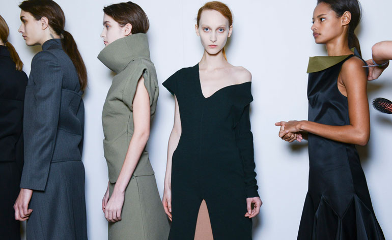 Four women, one in a blue dress, one in a grey dress, one in a black dress, and one in a black dress with an olive-green collar