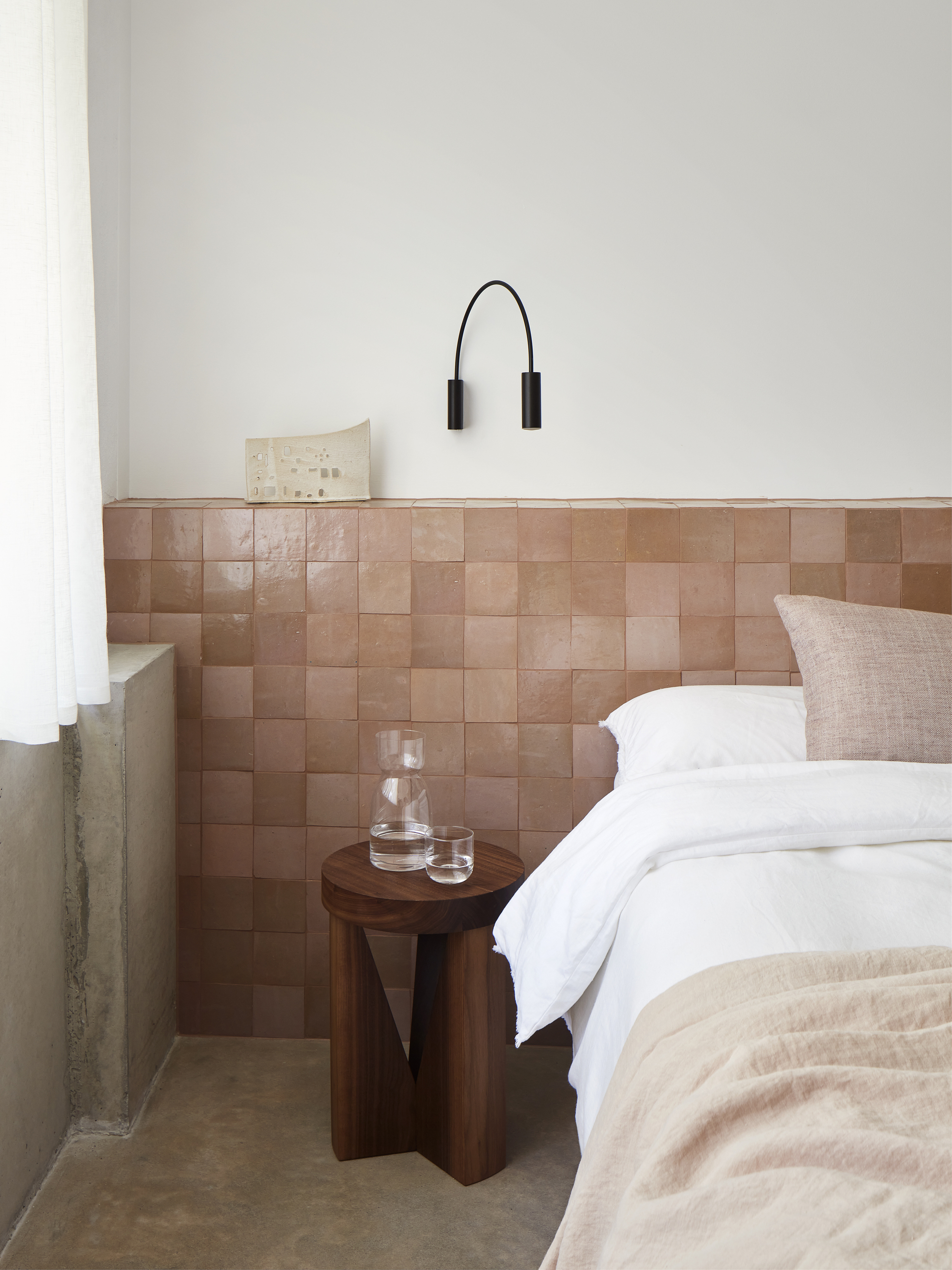 Image of a bedroom with an extended headboard made of tonal blush pink zellige tiles. The bedding is white and light pink and there is a wooden side table.