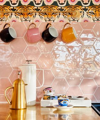 A kitchen countertop with pink tiles, mugs, a white kettle, and a gold French press