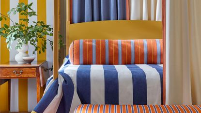 colorful bedroom with blue and white striped bedding, orange striped bolster pillow, striped bench at the end of the bed, curtains behind bed, yellow and white striped wallpaper, timber side table with vase with foliage in it