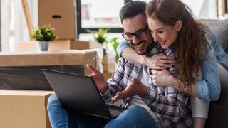 A couple sitting on a couch (a man and a woman), with the man holding a laptop and they&#039;re both looking at it