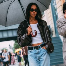 Laura Harrier is walking in the rain carrying an umbrella. She is wearing jeans, white tank top, black bomber, and black sunglasses. 