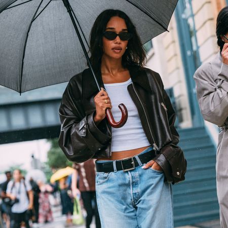 Laura Harrier is walking in the rain carrying an umbrella. She is wearing jeans, white tank top, black bomber, and black sunglasses. 